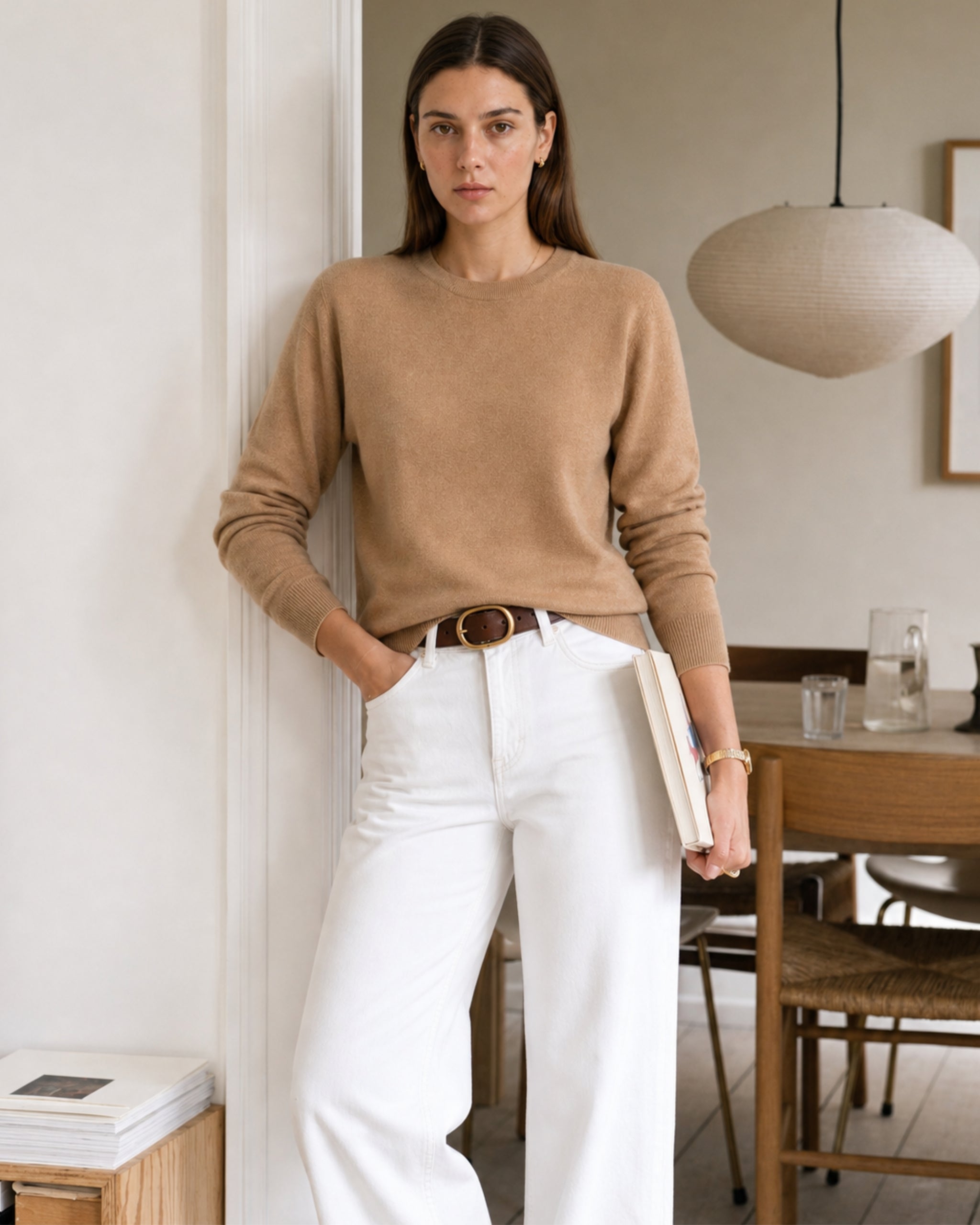 Woman in a camel merino sweater and white jeans holding a book at home